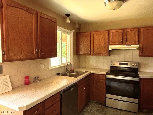 Kitchen with stainless steel appliances, tile countertops, under cabinet range hood, decorative backsplash, and brown cabinetry