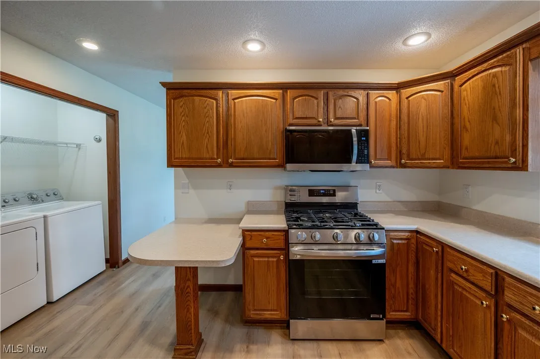 Kitchen with washing machine and clothes dryer, appliances with stainless steel finishes, light wood-style floors, light countertops, and brown cabinets
