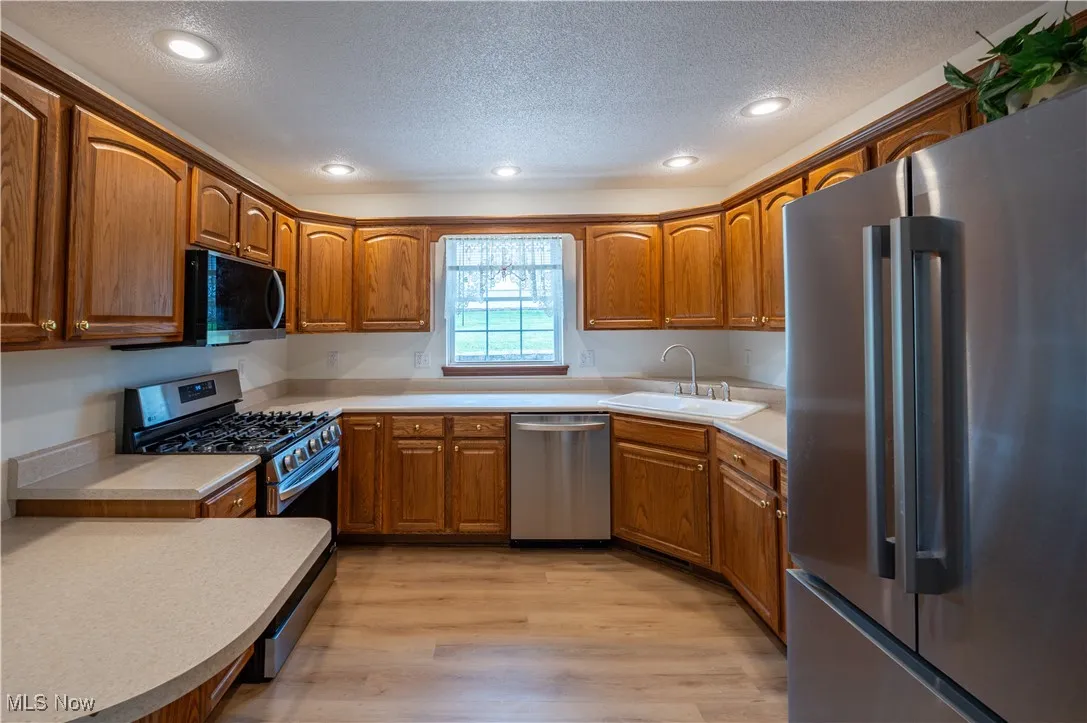 Kitchen with stainless steel appliances, a textured ceiling, brown cabinetry, light wood-type flooring, and recessed lighting