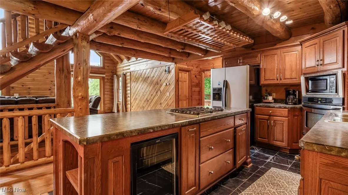 Kitchen featuring beverage cooler, wooden walls, stainless steel appliances, a wooden ceiling with exposed beams, and dark countertops