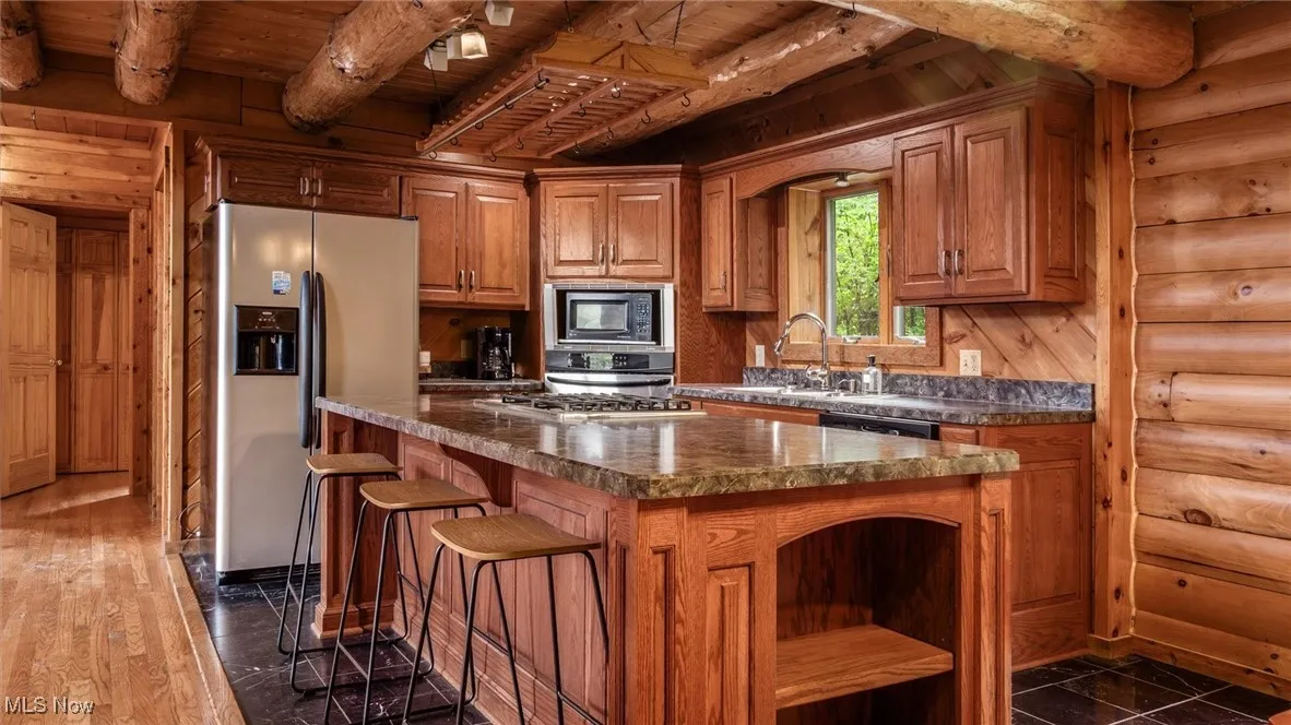 Kitchen featuring a wood ceiling with exposed beams, appliances with stainless steel finishes, rustic walls, a kitchen island, and open shelves