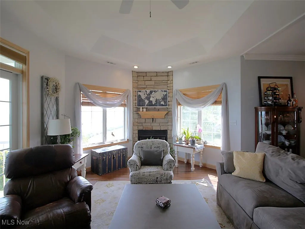 Living room with a raised ceiling, wood finished floors, ceiling fan, a fireplace, surrounding bay windows with lake views