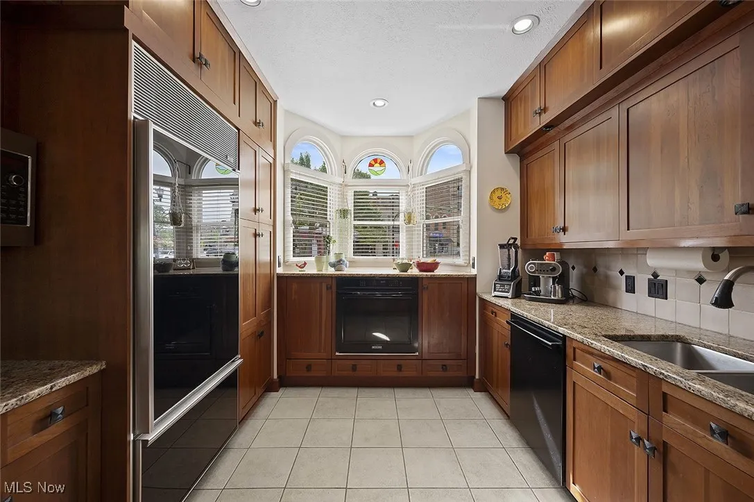 Kitchen featuring brown cabinets, backsplash, light stone countertops, black dishwasher, and light tile patterned floors