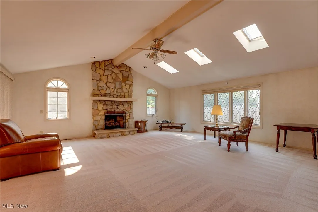 Carpeted living area with beamed ceiling, a skylight, a stone fireplace, ceiling fan, and high vaulted ceiling