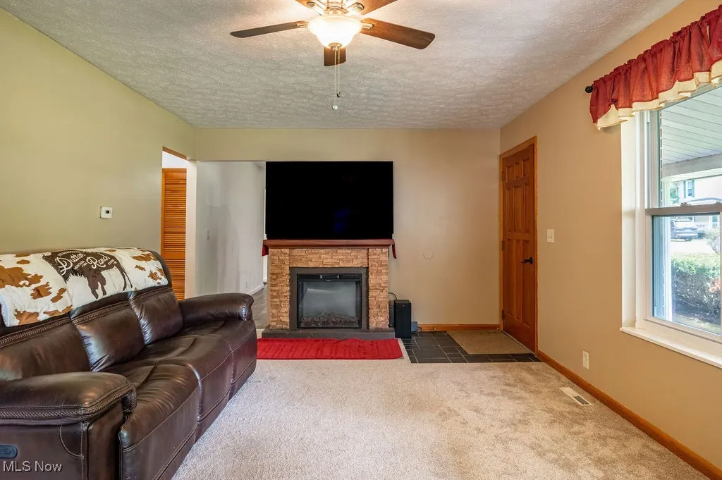 Carpeted living area with a textured ceiling, a ceiling fan, and a stone fireplace