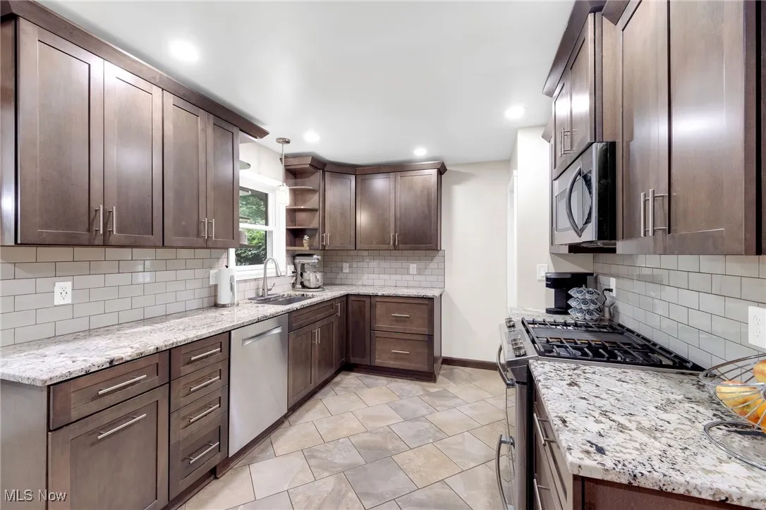 Kitchen with stainless steel appliances, open shelves, tasteful backsplash, light stone counters, and dark brown cabinets