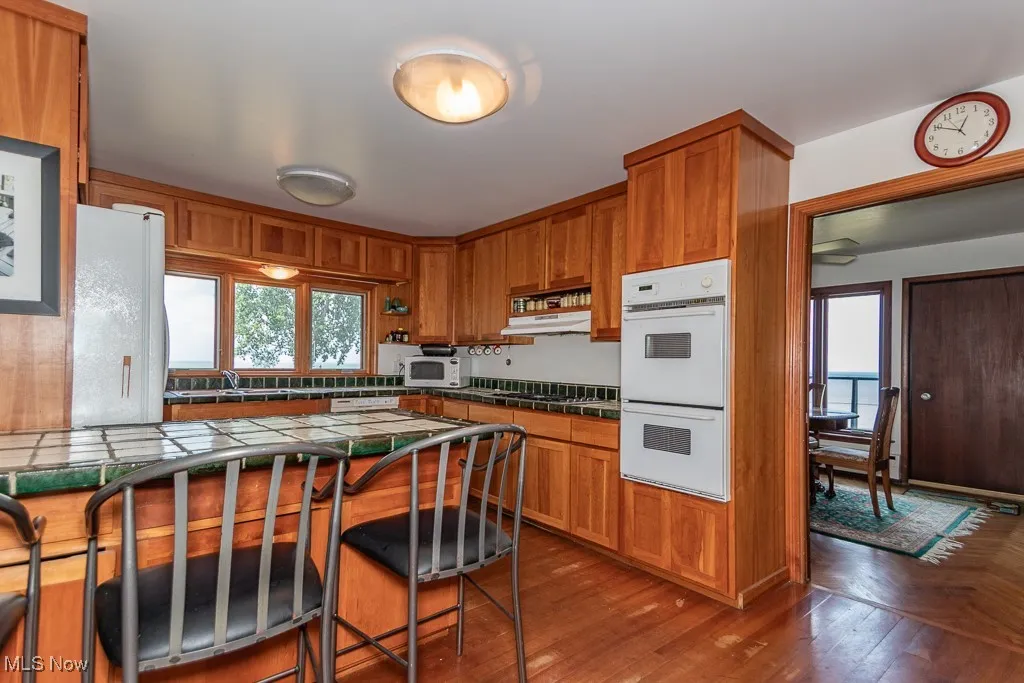 Kitchen with tile countertops, white appliances, brown cabinetry, a breakfast bar area, and under cabinet range hood