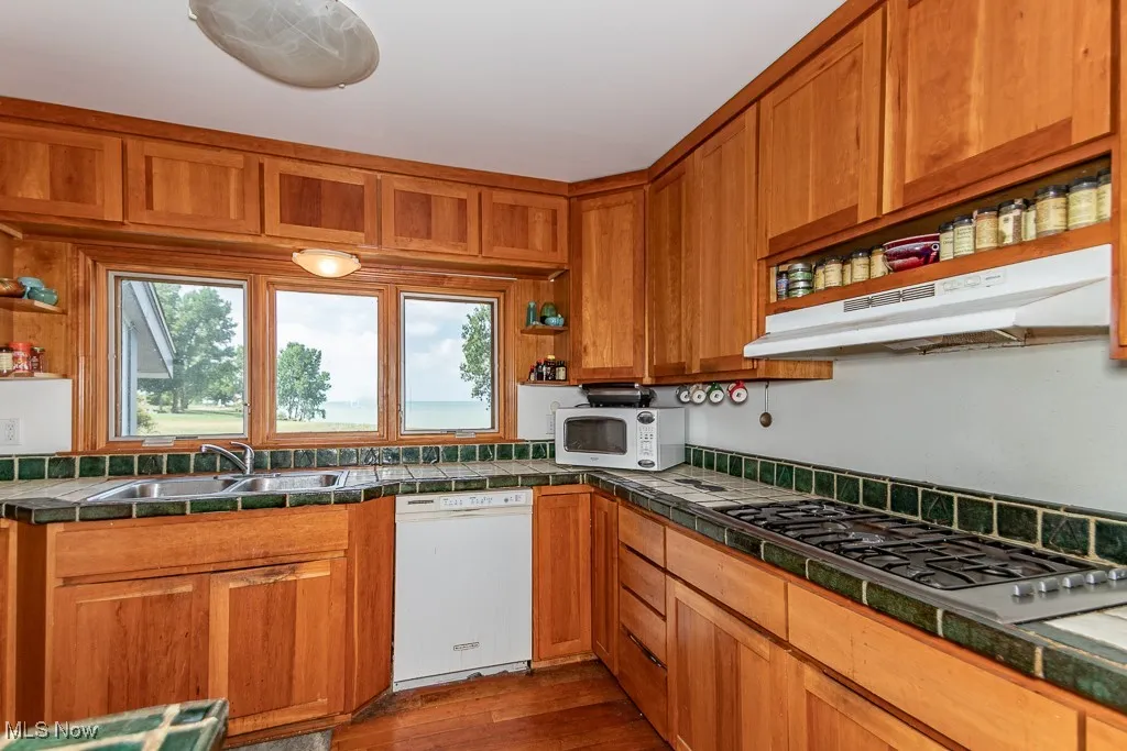 Kitchen featuring white appliances, brown cabinetry, under cabinet range hood, tile countertops, and dark wood finished floors