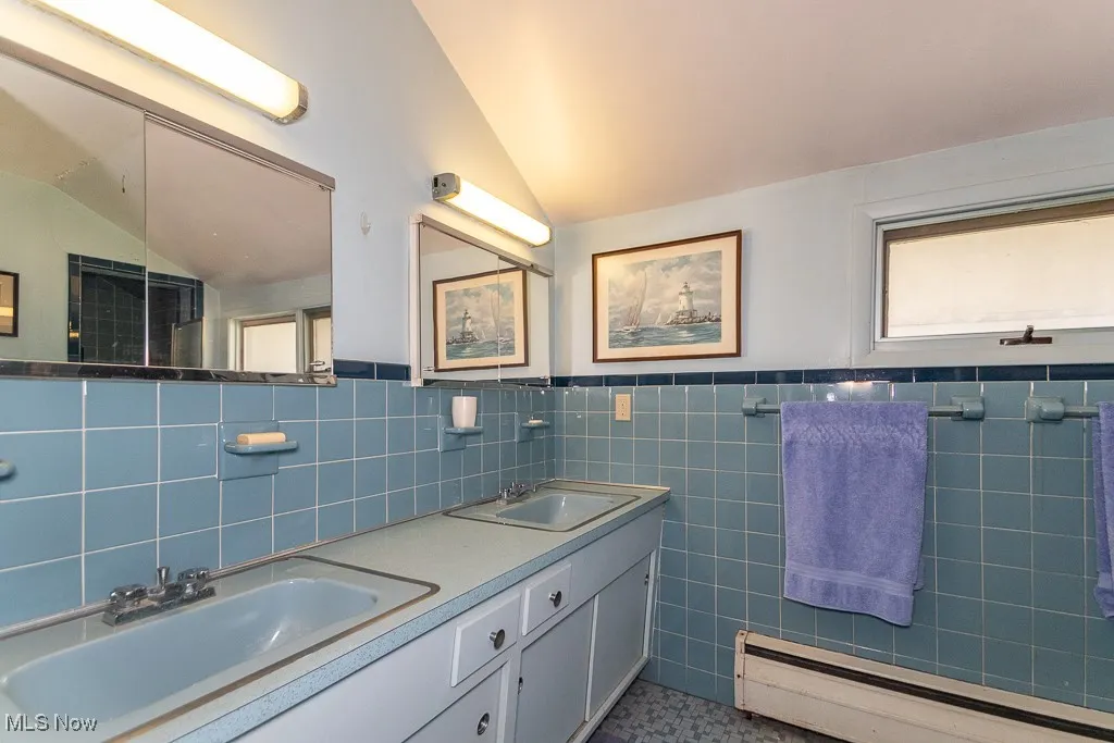 Full bath featuring tile walls, a baseboard radiator, lofted ceiling, double vanity, and wainscoting