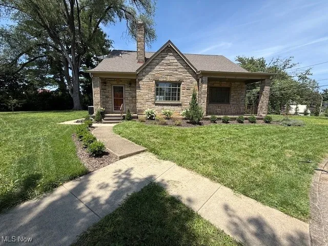 View of front facade with stone siding, a front yard, a chimney, a porch, and roof with shingles