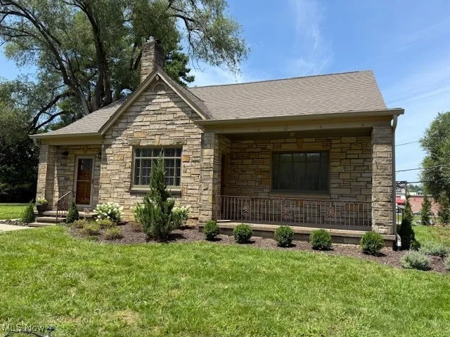 View of front of house featuring stone siding, a front yard, covered porch, a chimney, and a shingled roof