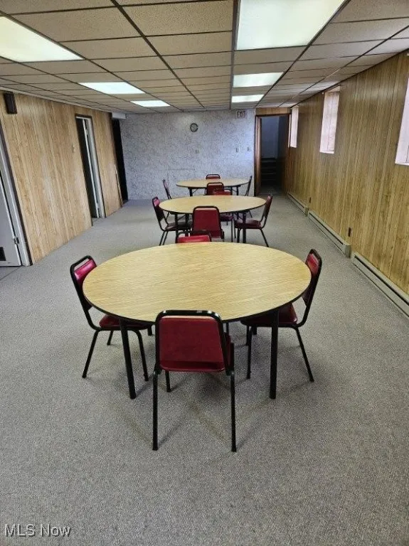 Dining area with light carpet, wooden walls, a drop ceiling, a baseboard radiator, and baseboard heating