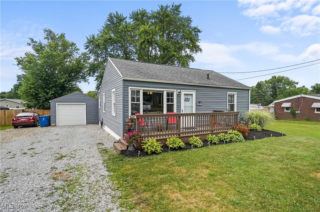 View of front facade with a wooden deck, gravel driveway, an outbuilding, a detached garage, and roof with shingles