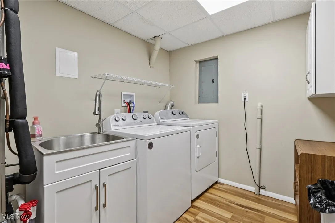 Laundry room featuring washing machine and clothes dryer, light wood finished floors, utility tub, electric panel, and cabinet space and compact attached ironing board.