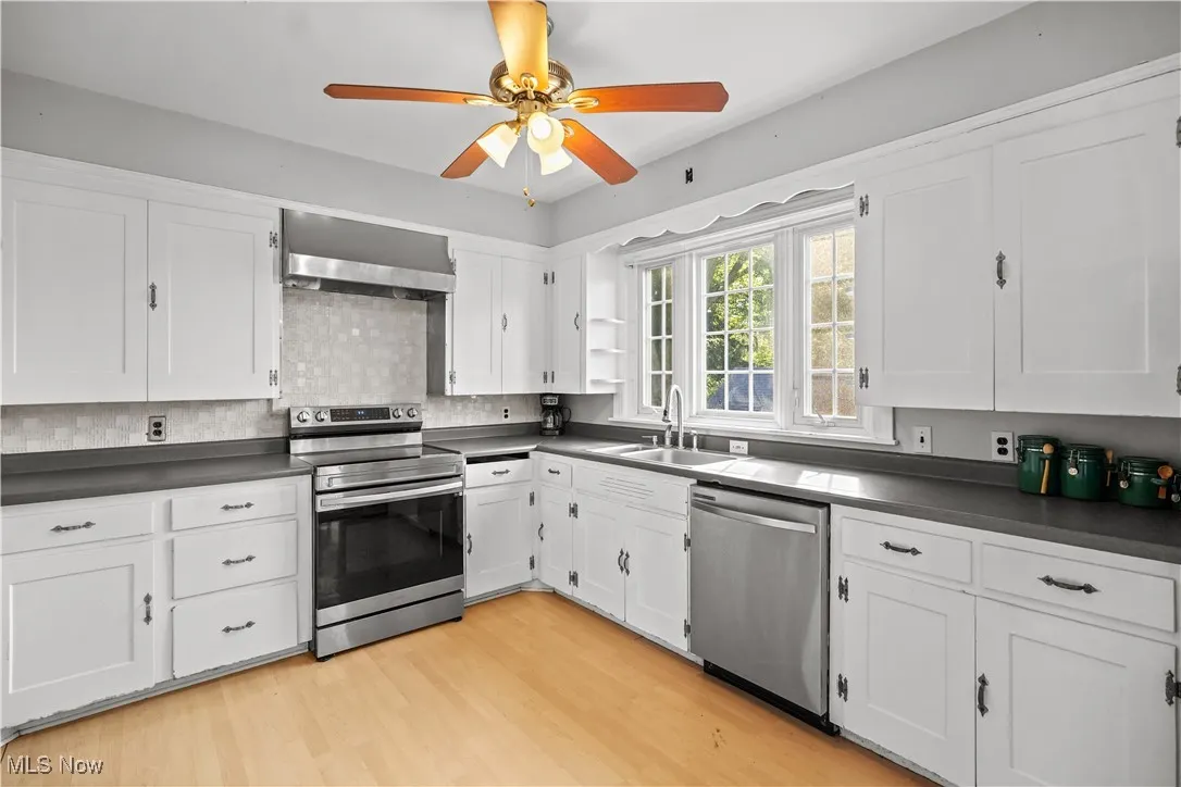 Kitchen with appliances with stainless steel finishes, dark countertops, wall chimney range hood, white cabinetry, and a ceiling fan