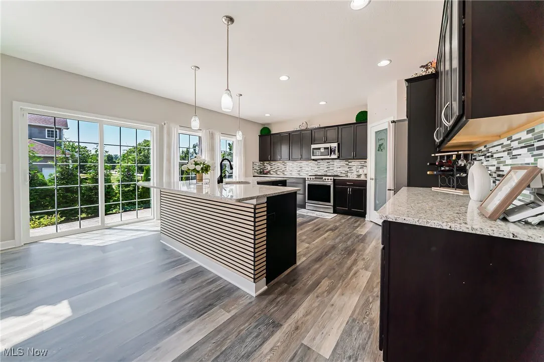Kitchen featuring backsplash, wood finished floors, appliances with stainless steel finishes, light stone countertops, and a center island with sink