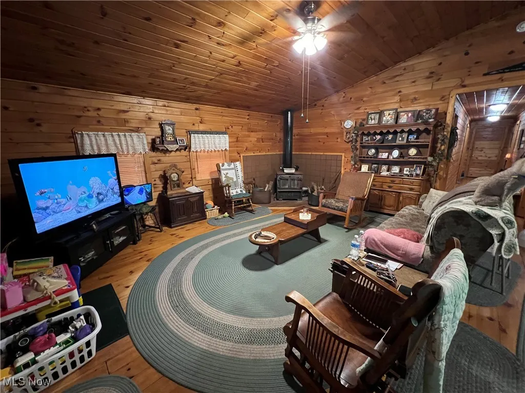 Living area with lofted ceiling, a wood stove, wooden walls, wood finished floors, and a ceiling fan