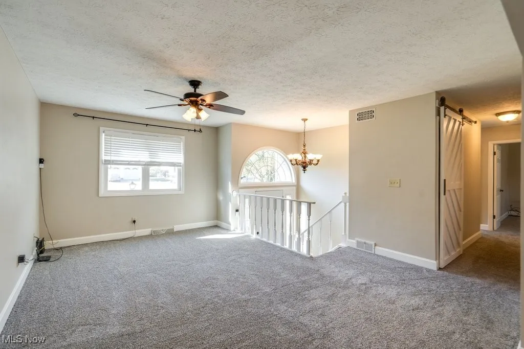 Carpeted spare room featuring a textured ceiling, ceiling fan, a barn door, and a chandelier