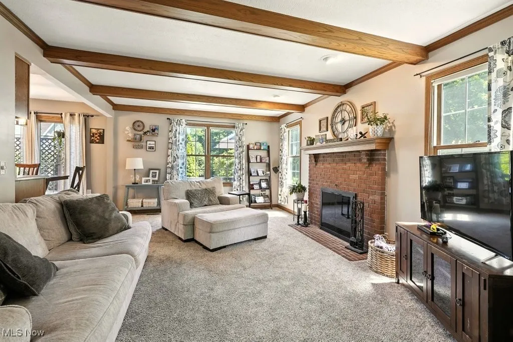 Carpeted living area with healthy amount of natural light, a brick fireplace, beam ceiling, and crown molding