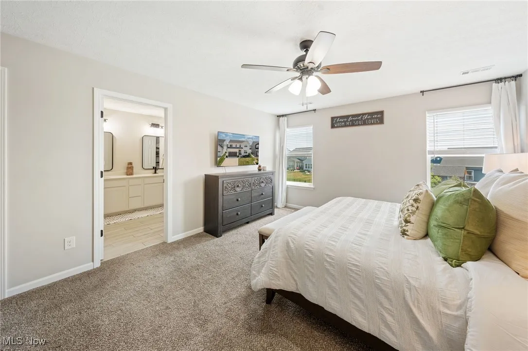 Carpeted bedroom featuring multiple windows, ensuite bath, and a ceiling fan