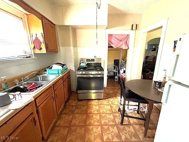 Kitchen with freestanding refrigerator, gas stove, brown cabinetry, light floors, and light countertops