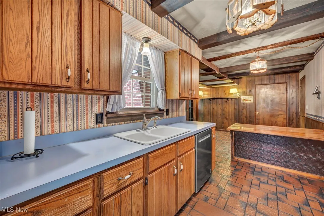 Kitchen with wallpapered walls, beam ceiling, light countertops, brown cabinets, and dishwasher