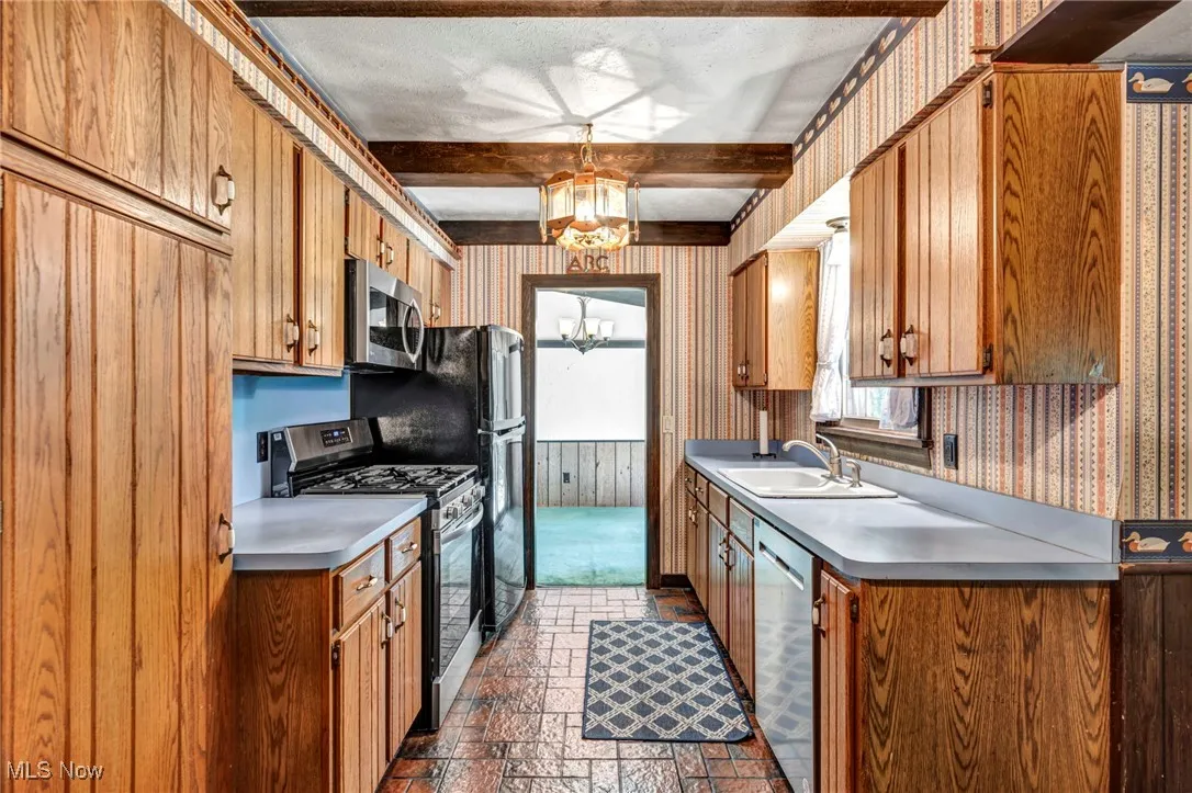 Kitchen with wallpapered walls, appliances with stainless steel finishes, beamed ceiling, a chandelier, and brick patterned floors
