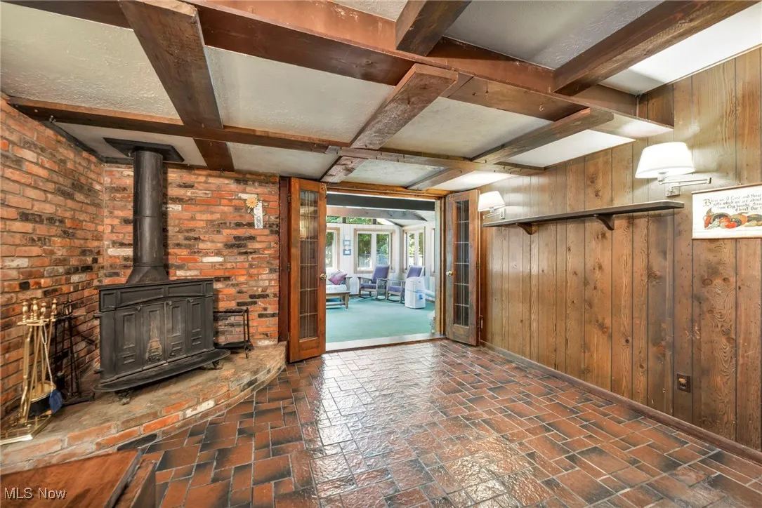 Unfurnished living room featuring beam ceiling, coffered ceiling, wooden walls, and a wood stove