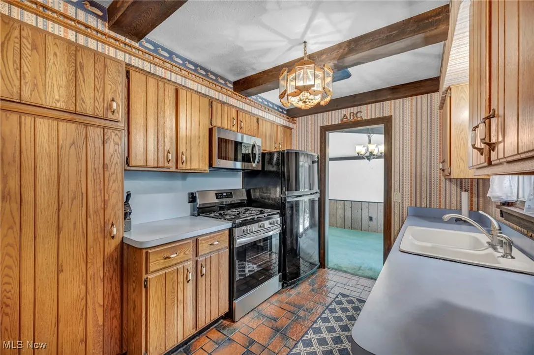 Kitchen with a chandelier, beamed ceiling, wallpapered walls, appliances with stainless steel finishes, and light countertops