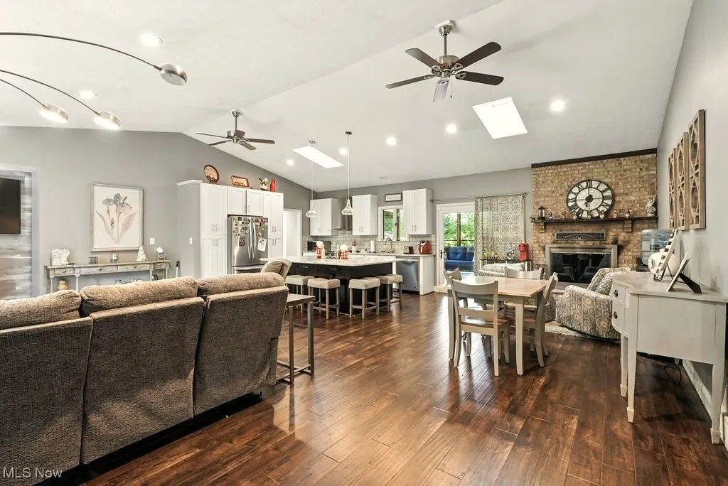 Living room featuring ceiling fan, vaulted ceiling, a brick fireplace, a skylight, and dark wood finished floors
