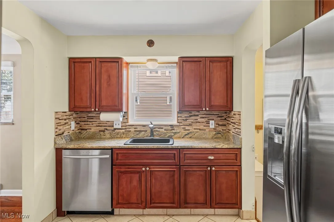 Kitchen with dark brown cabinets, stainless steel appliances, backsplash, arched walkways, and light stone counters