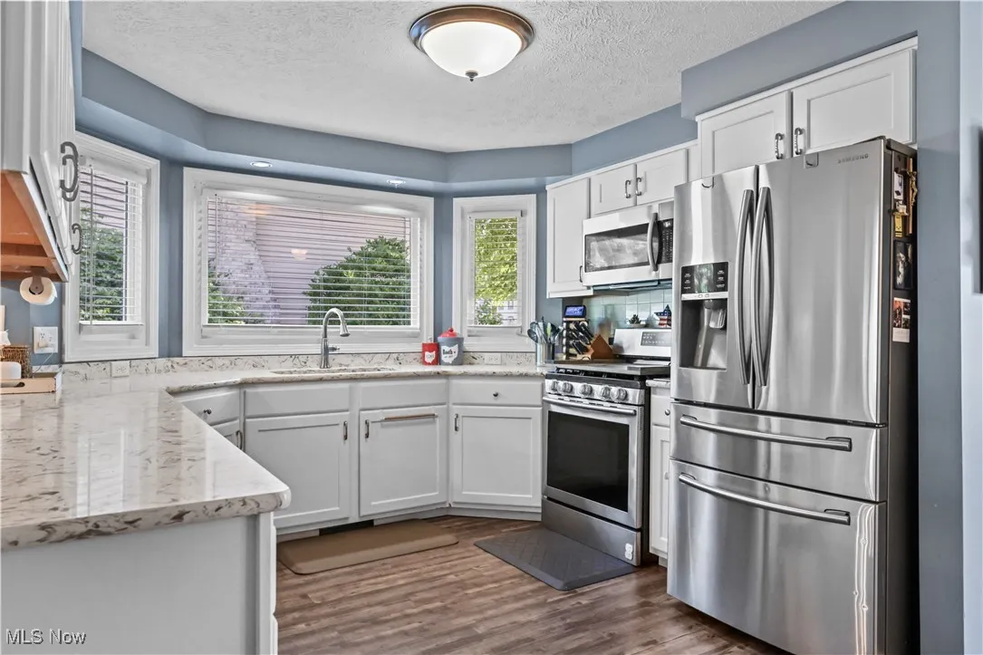 Kitchen with stainless steel appliances, a textured ceiling, dark wood-style floors, white cabinets, and light stone counters