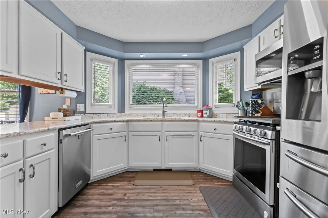 Kitchen with appliances with stainless steel finishes, a textured ceiling, white cabinets, dark wood-type flooring, and light stone counters