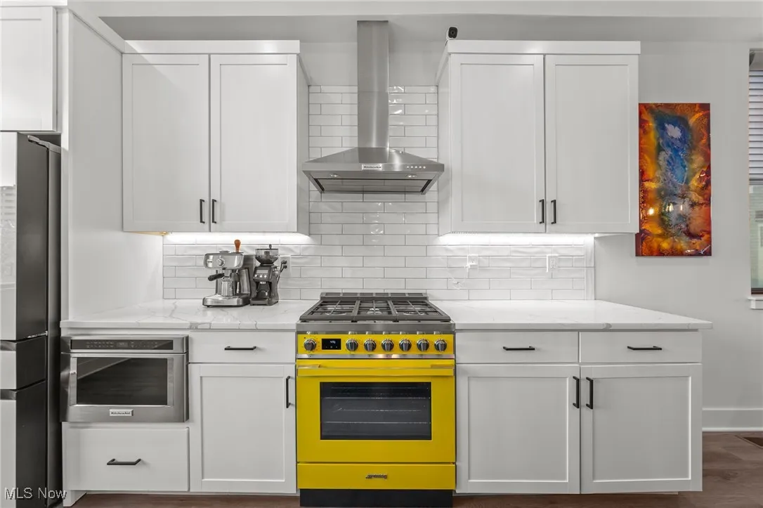 Kitchen featuring stainless steel appliances, wall chimney range hood, white cabinets, and light stone countertops