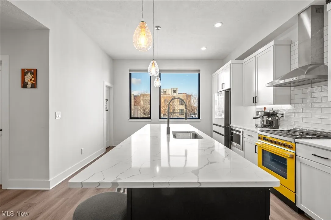 Kitchen featuring wall chimney exhaust hood, light wood-style flooring, gas range oven, tasteful backsplash, and an island with sink
