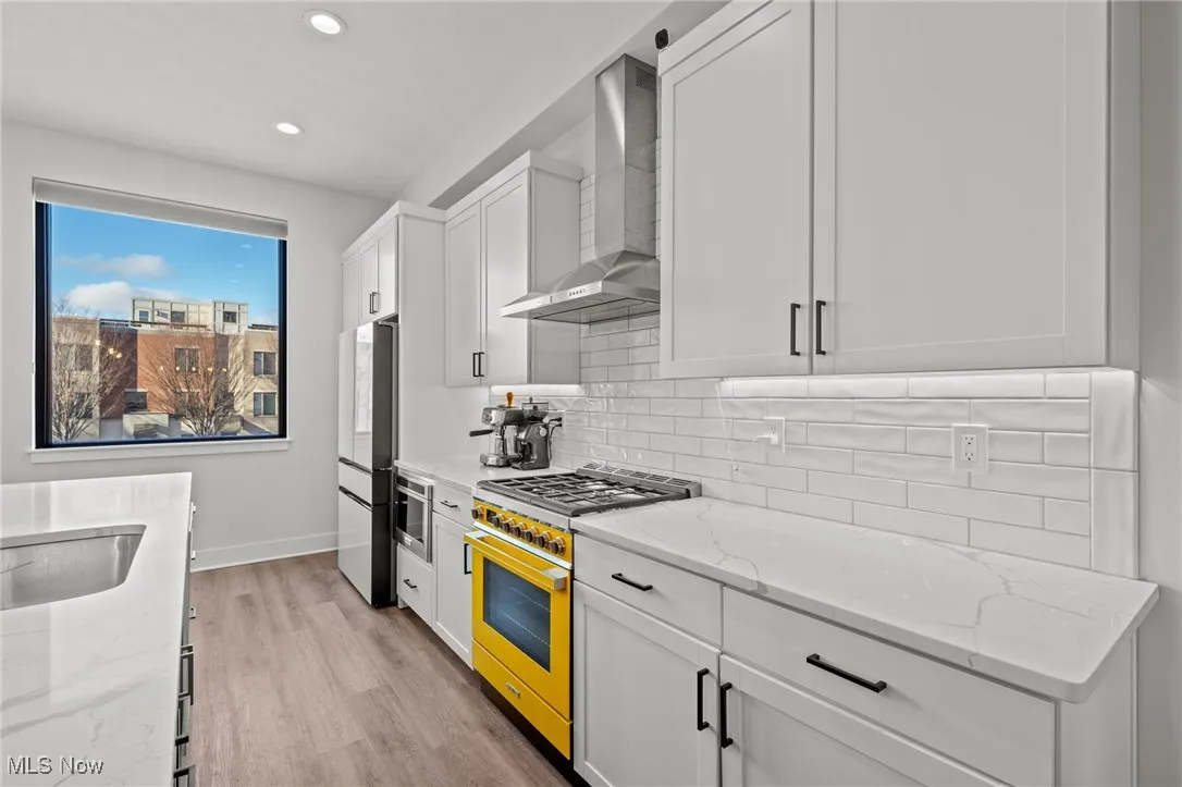 Kitchen featuring wall chimney exhaust hood, gas stove, white cabinets, decorative backsplash, and light wood-type flooring