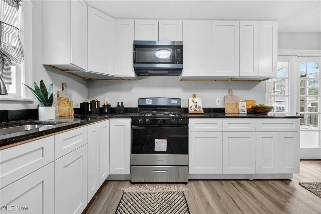 Kitchen with gas range, white cabinets, stainless steel microwave, light wood-style floors, and dark stone countertops