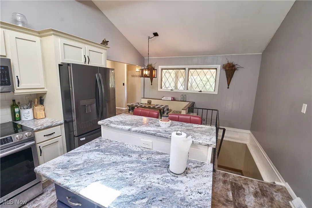Kitchen featuring appliances with stainless steel finishes, vaulted ceiling, a kitchen island, a chandelier, and light stone counters