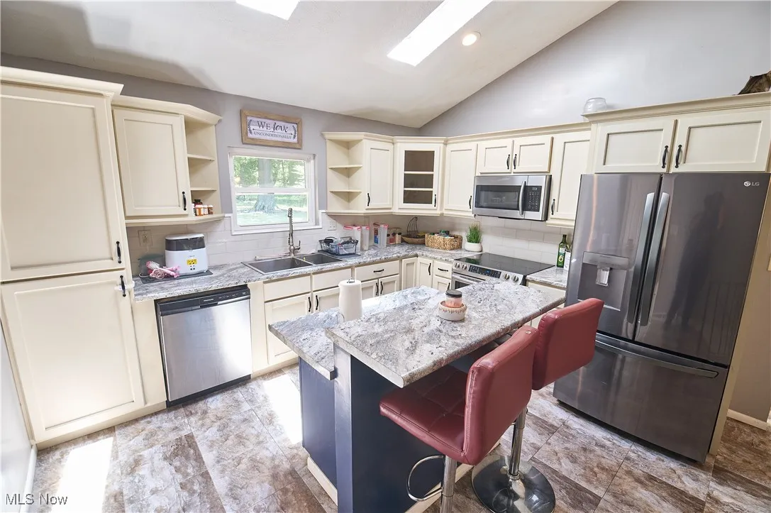 Kitchen featuring open shelves, appliances with stainless steel finishes, vaulted ceiling, a kitchen island, and light stone countertops