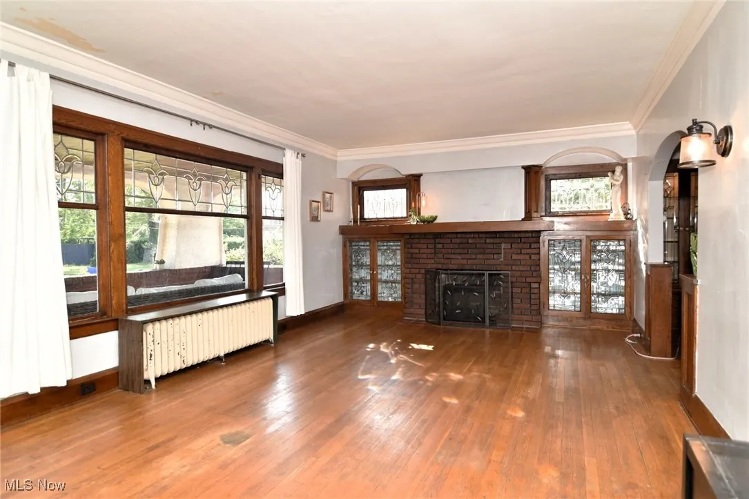 Unfurnished living room with radiator, wood finished floors, healthy amount of natural light, a brick fireplace, and crown molding