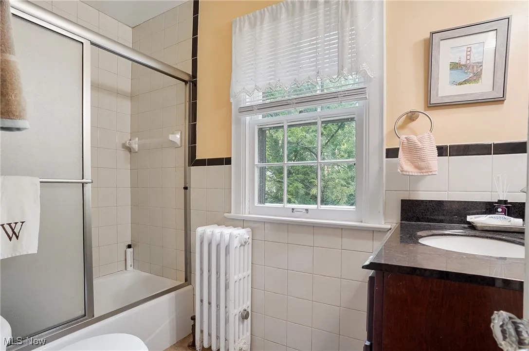 Bathroom featuring radiator, tile walls, vanity, shower / bath combination with glass door, and wainscoting