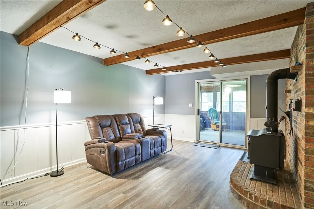 Living area featuring a wood stove, beamed ceiling, wainscoting, vinyl plank floors and sun room access