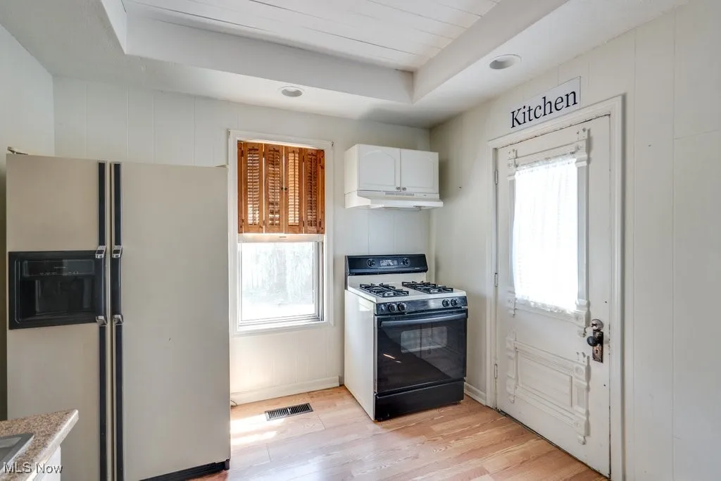 Kitchen with white fridge with ice dispenser, black gas range oven, light wood finished floors, white cabinetry, and under cabinet range hood