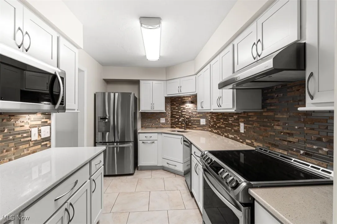 Kitchen with appliances with stainless steel finishes, tasteful backsplash, under cabinet range hood, light tile patterned floors, and light stone countertops