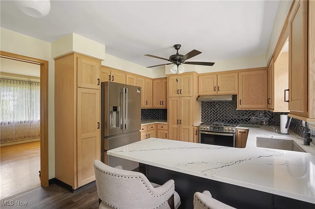 Kitchen featuring a peninsula, appliances with stainless steel finishes, decorative backsplash, a ceiling fan, and dark wood-style floors