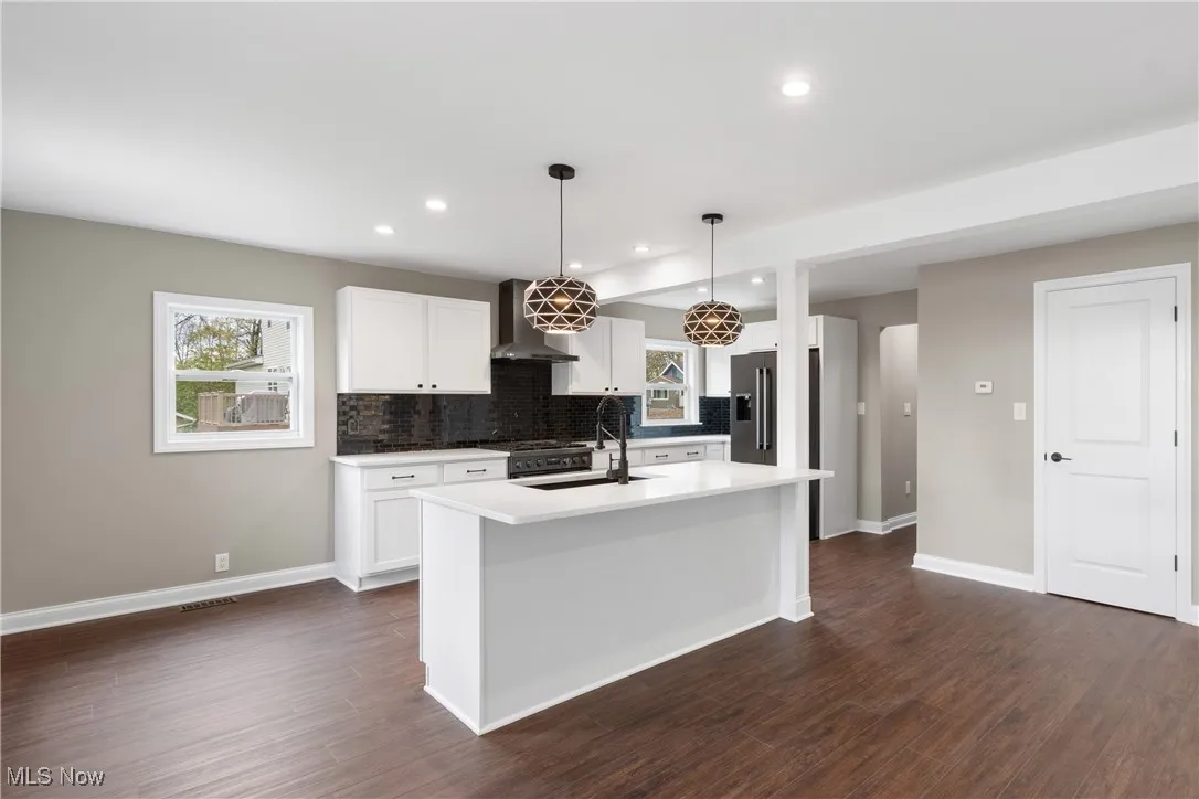 Kitchen featuring dark wood-type flooring, wall chimney exhaust hood, white cabinetry, a center island with sink, and recessed lighting