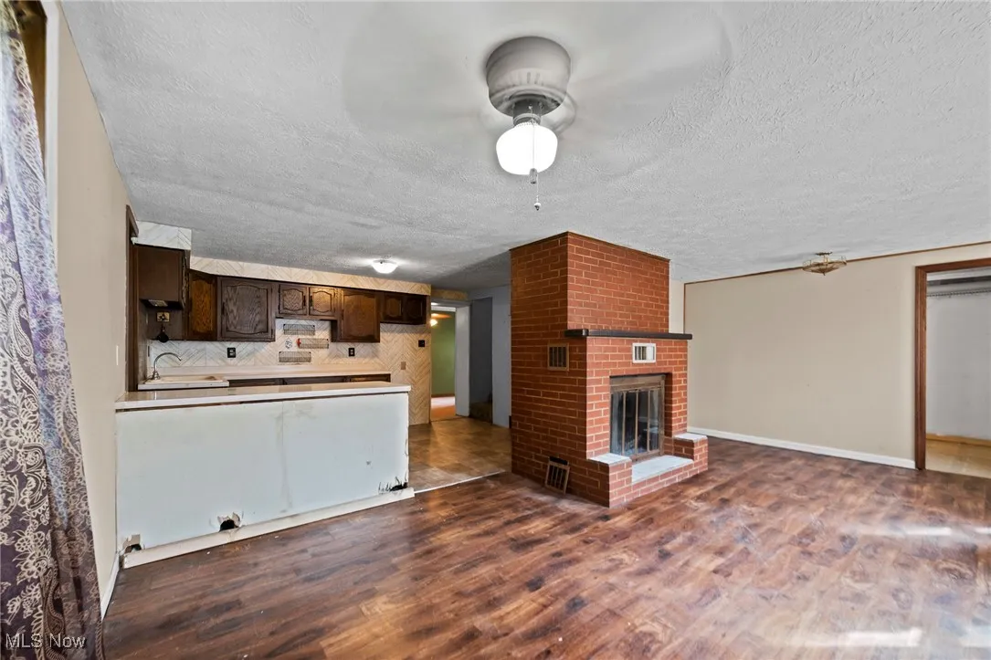 Unfurnished living room featuring a ceiling fan, a textured ceiling, dark wood-style flooring, and a brick fireplace