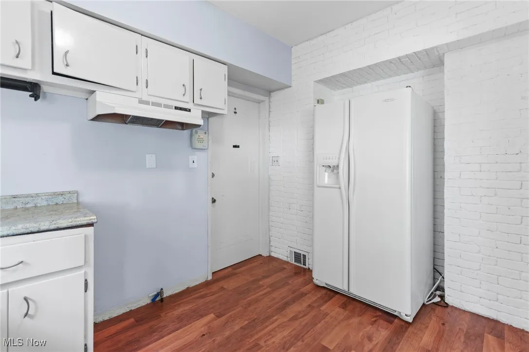 Kitchen with brick wall, white refrigerator with ice dispenser, dark wood-type flooring, light countertops, and white cabinets