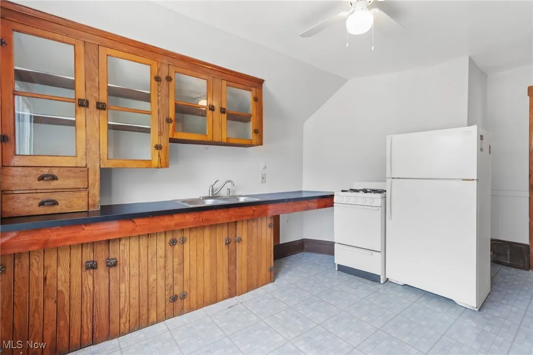 Kitchen with white appliances, brown cabinets, a ceiling fan, glass insert cabinets, and vaulted ceiling