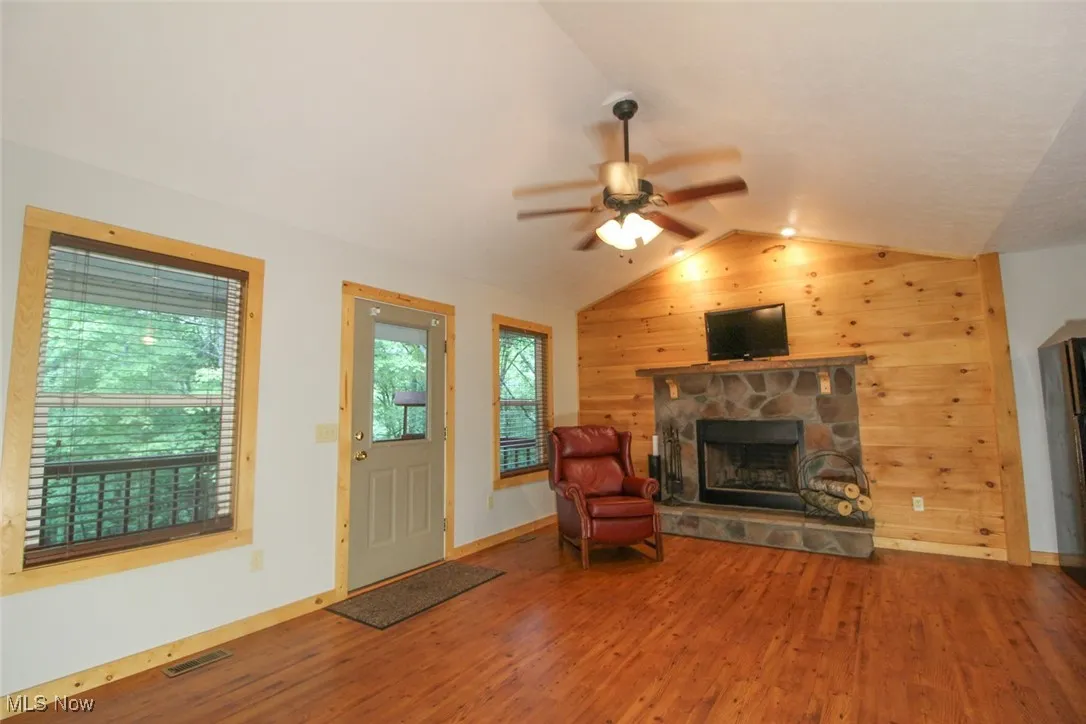 Living room with vaulted ceiling, wooden walls, wood finished floors, ceiling fan, and a fireplace